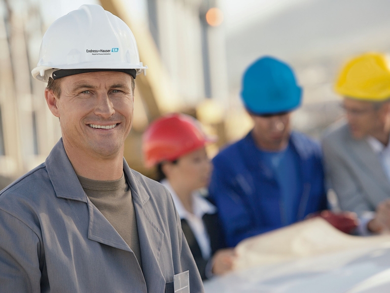 Con Endress+Hauser tiene un socio fiable para los servicios de ingeniería Un ingeniero de Endress+Hauser sonríe a la cámara, otros ingenieros están detrás de él